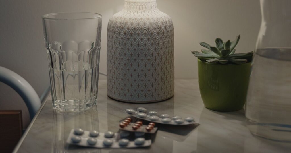 Mesa de cabeceira com copo de água, cartelas de comprimidos e a base de um abajur aceso: a rotina do remédio oral tomado em casa, como a enzalutamida (Xtandi) pedida ao plano de saúde